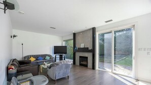 Sala de estar moderna com sofá, poltrona, televisão, lareira e grandes portas de vidro deslizantes que dão para um pátio.