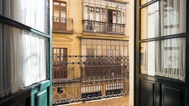 Blick durch ein Fenster mit einem Metallbalkonläufer, einem Backboden und einem Gebäude mit mehreren Fenstern und Balkonen im Hintergrund.