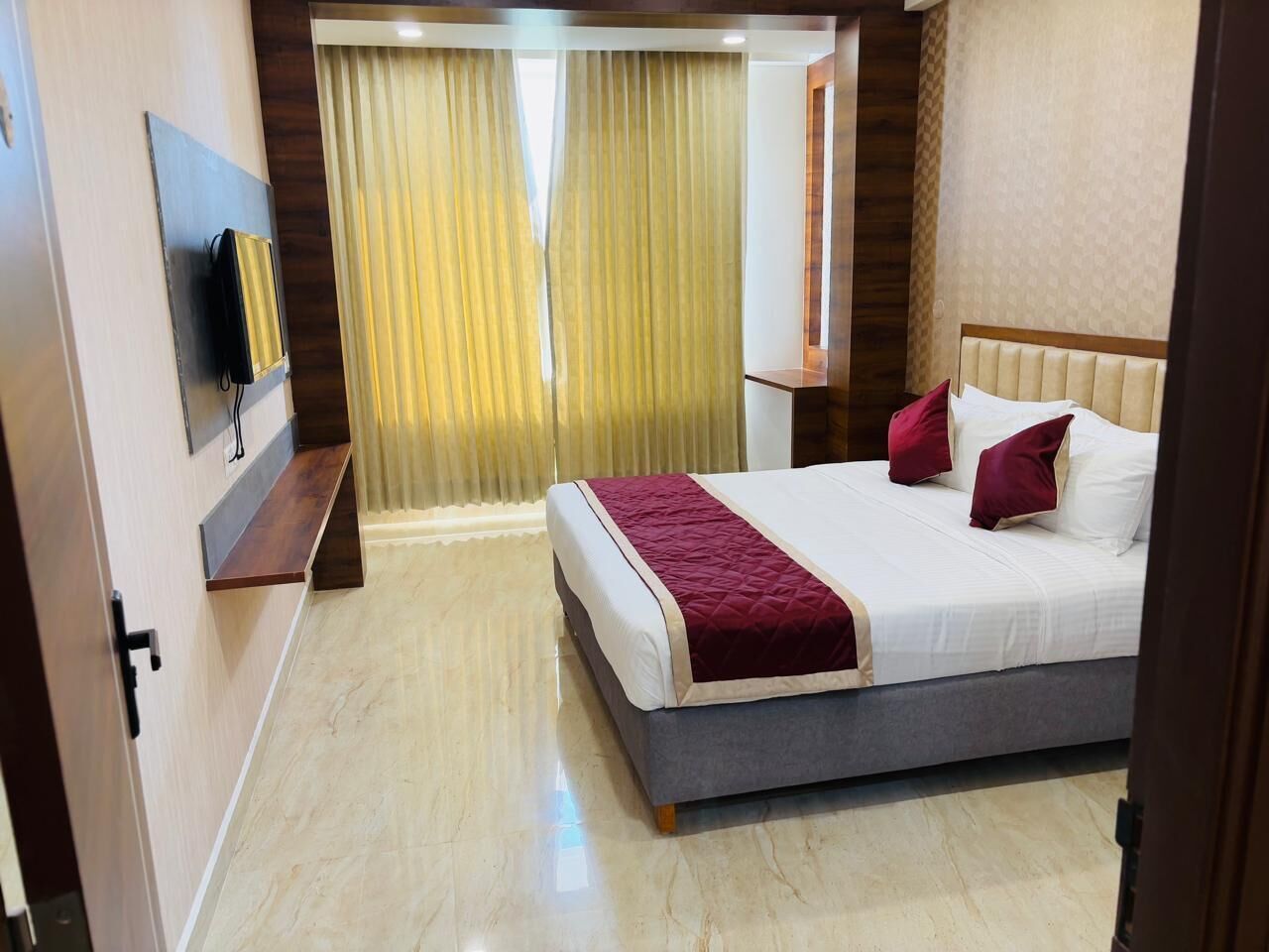 A hotel room with a bed, a TV mounted on the wall, a bedside table, and a window with curtains.