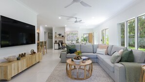 A spacious living room with a large flat-screen TV, a grey sectional sofa, a wooden coffee table, and a ceiling fan. - Sorrento Breeze | Port Douglas (PORT DOUGLAS)