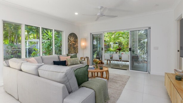 A spacious living room with a large sofa, a coffee table, and a dining area with chairs. There are large windows providing a view of greenery outside. - Sorrento Breeze | Port Douglas (PORT DOUGLAS)