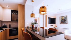 A modern kitchen with wooden cabinets, a black countertop, and a central island with chairs. There is a dining area with a white sofa and a painting on the wall. - Belgrade Rooftop Experience, Modern Luxury & Views (Beograd)