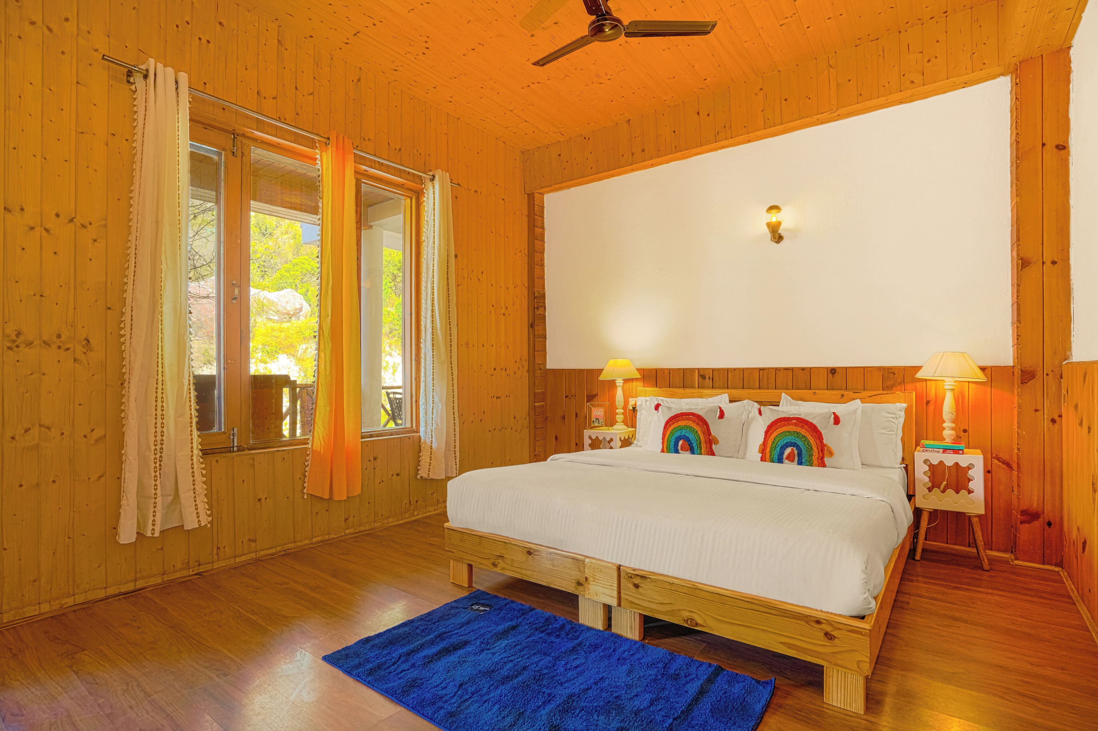 Quarto com painéis de madeira, cama, duas luminárias de cabeceira, ventoinha no teto e janela com cortinas.