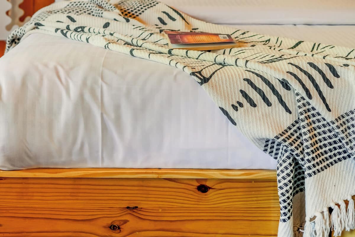 A neatly made bed with white bedding and rainbow-patterned pillows, a wooden headboard, and a bedside table with a lamp and a framed picture.