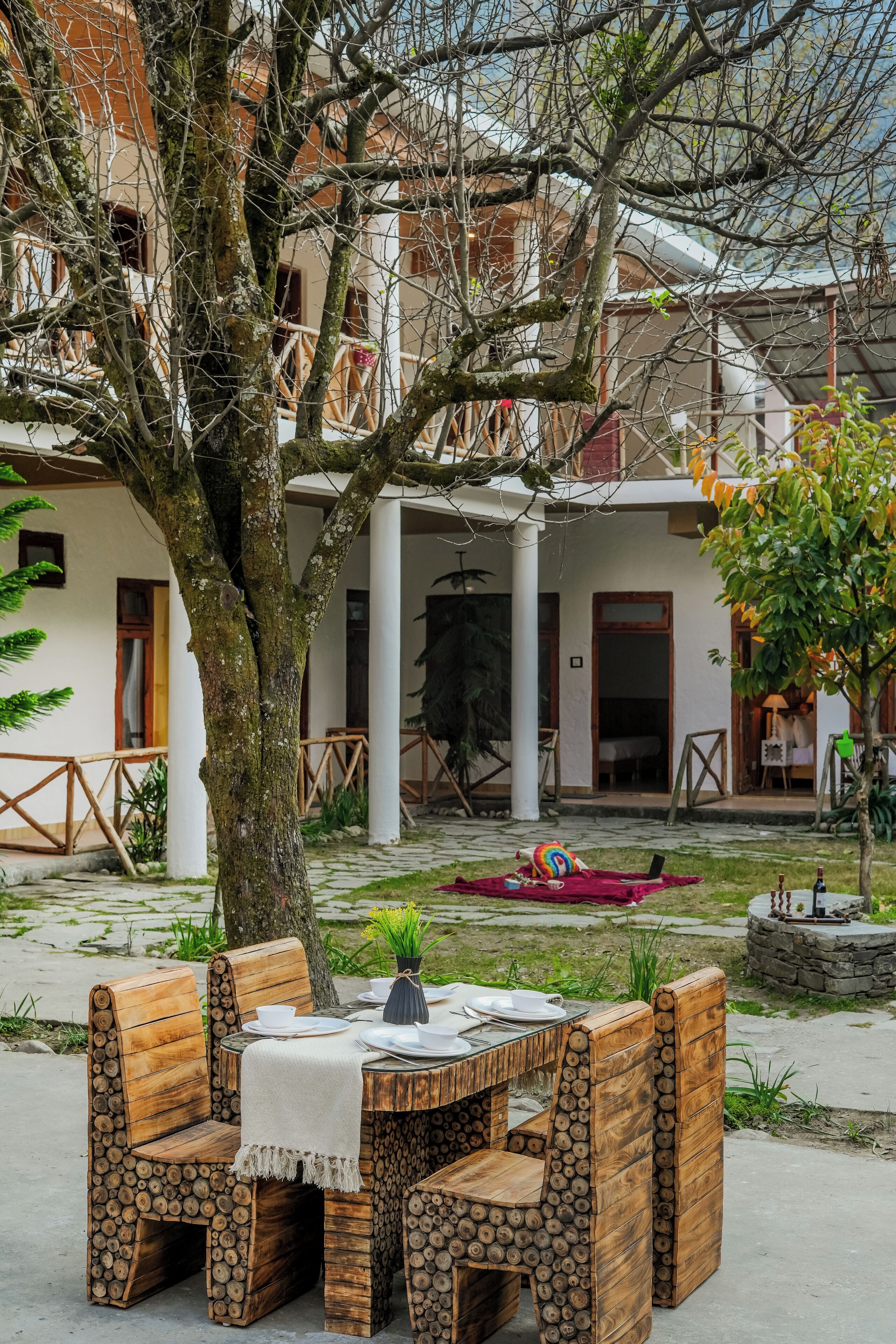 A rustic outdoor seating area with wooden log chairs and a table, surrounded by a garden and a building with balconies.
