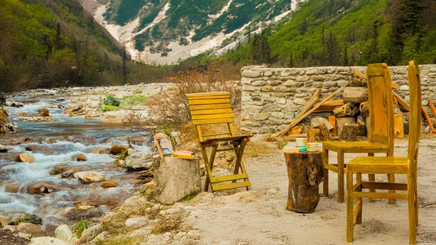 Ein rustikaler Sitzbereich im Freien mit Holzkufen und -tischen, einer Steinsäule und einem Fluss im Hintergrund.