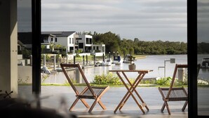 Une vue prise de l’intérieur d’un bâtiment, donnant sur une étendue d’eau où se trouvent des bateaux, ainsi que sur deux chaises en bois et une petite table.