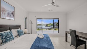 Une chambre à coucher comprenant un lit, un bureau, une chaise, un ventilateur de plafond, une fenêtre donnant sur un plan d’eau et de la verdure, ainsi qu’un tableau accroché au mur.
