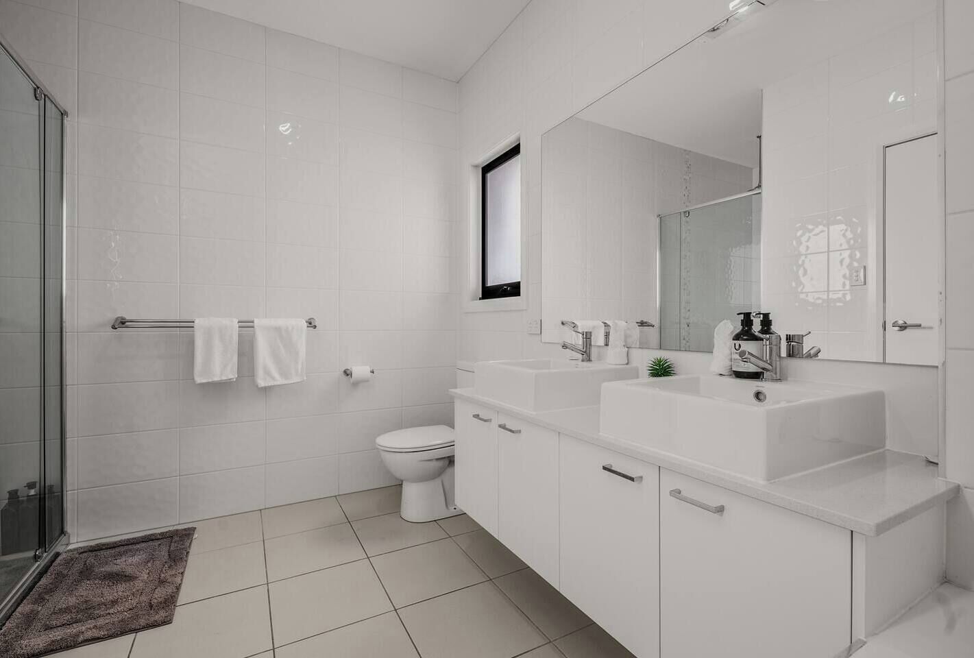 Ein modernes Badezimmer mit Duschabtrennung aus Glas, weißen Fliesen, einer Toilette, einem Waschbecken mit Spiegel und einer kleinen Pflanze.