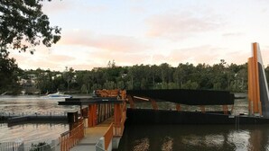 Puente sobre un río con sendero para peatones, rodeado de árboles y un paisaje urbano distante.