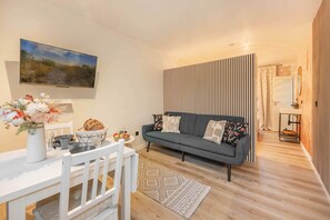 Un salon moderne avec un téléviseur à écran plat, un canapé gris orné de coussins à motifs, une table à manger blanche avec des chaises et un plancher en bois.