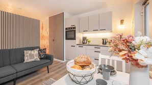 Un salon moderne avec un canapé, une table à manger agrémentée d’un panier à pain, et un coin cuisine équipé de placards et d’une machine à café.