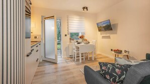 Une cuisine moderne avec des meubles blancs, un coin repas comprenant une table et des chaises, un téléviseur fixé au mur et une vue sur l’extérieur par une porte.