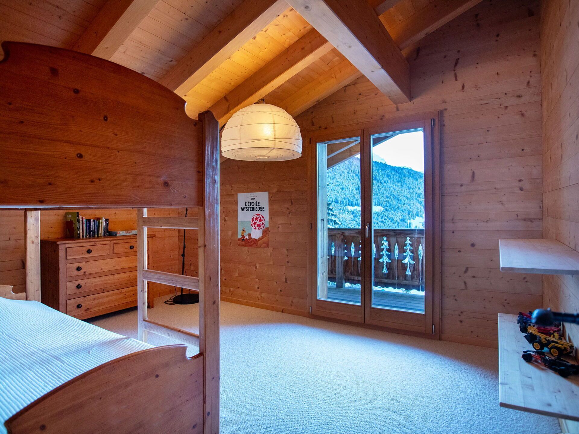 A wooden cabin room with a bunk bed, a dresser, a desk, and a large window overlooking a snowy mountain.