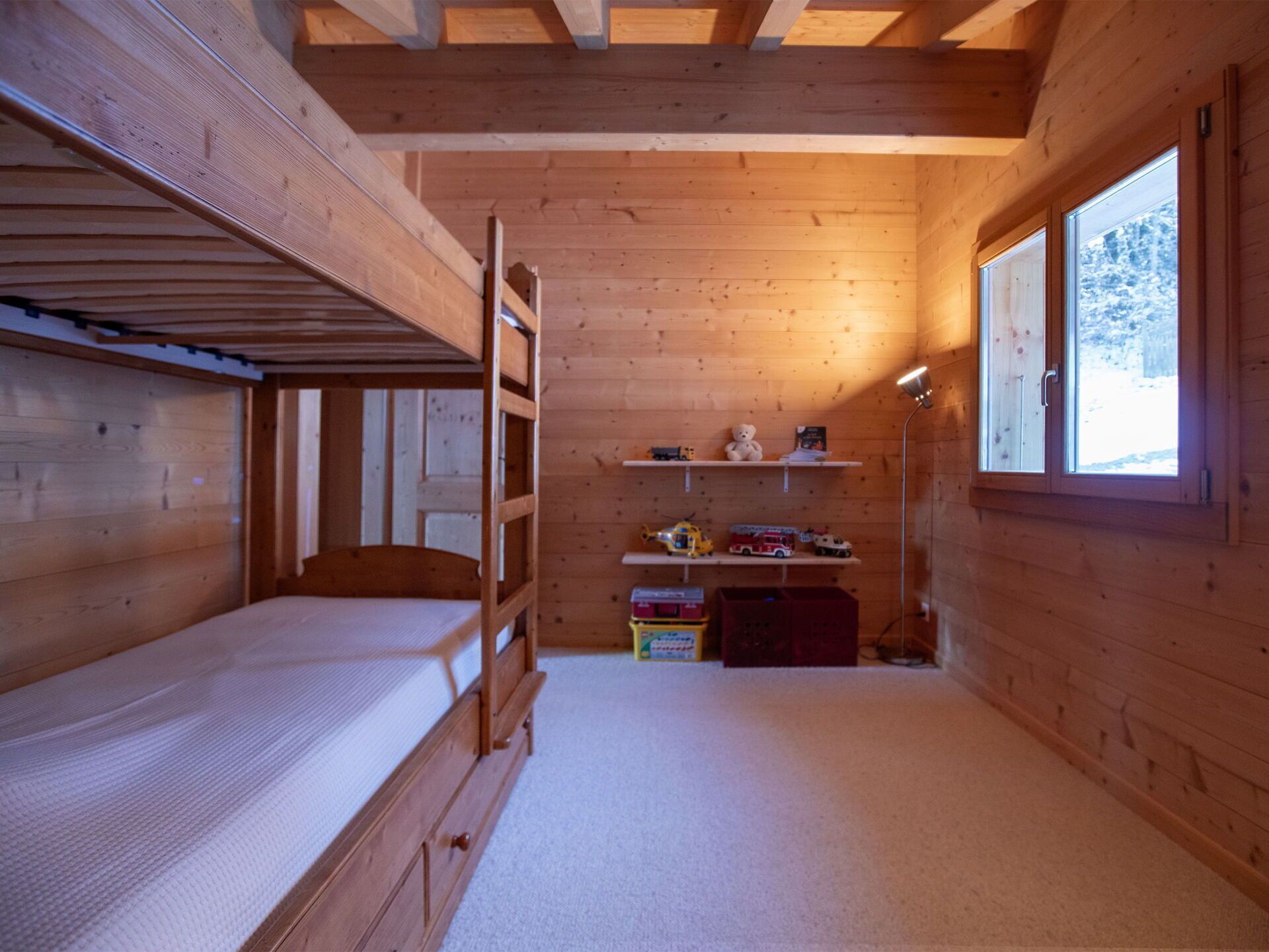 Quarto de cabana de madeira com beliches, uma escrivaninha, prateleiras com brinquedos e uma janela.