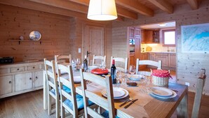 A wooden interior with a dining table set for four, wooden chairs, a hanging light fixture, and a kitchen visible through an open door. - Wonderful private villa for 8 guests with terrace (Alpe Des Chaux)