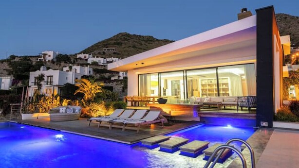 Uma villa moderna com piscina, espreguiçadeiras e vista para a colina ao redor.