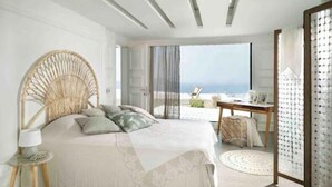 Quarto moderno com cama grande, mesa lateral redonda, cadeira de vime e vista para o mar.