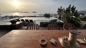 Mesa de madeira com cadeiras e uma tigela, com vista para uma piscina e uma paisagem cênica do mar e ilhas ao longe.