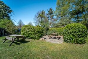 Un área de picnic con mesas y bancos de madera, rodeada de setos y árboles bien cuidados.