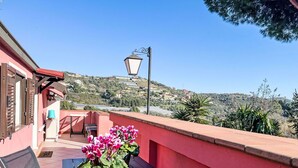 Un balcón con mesa y sillas, una planta en maceta con flores y vistas a una colina con edificios.