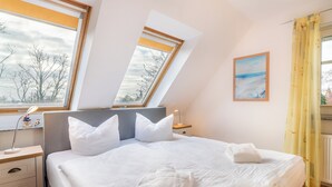 A neatly made bed with white linens and pillows, a bedside table with a lamp, and a window with a view of trees and a cloudy sky. - House Dünenwald 11 - small, cozy domicile with balcony (Zinnowitz)