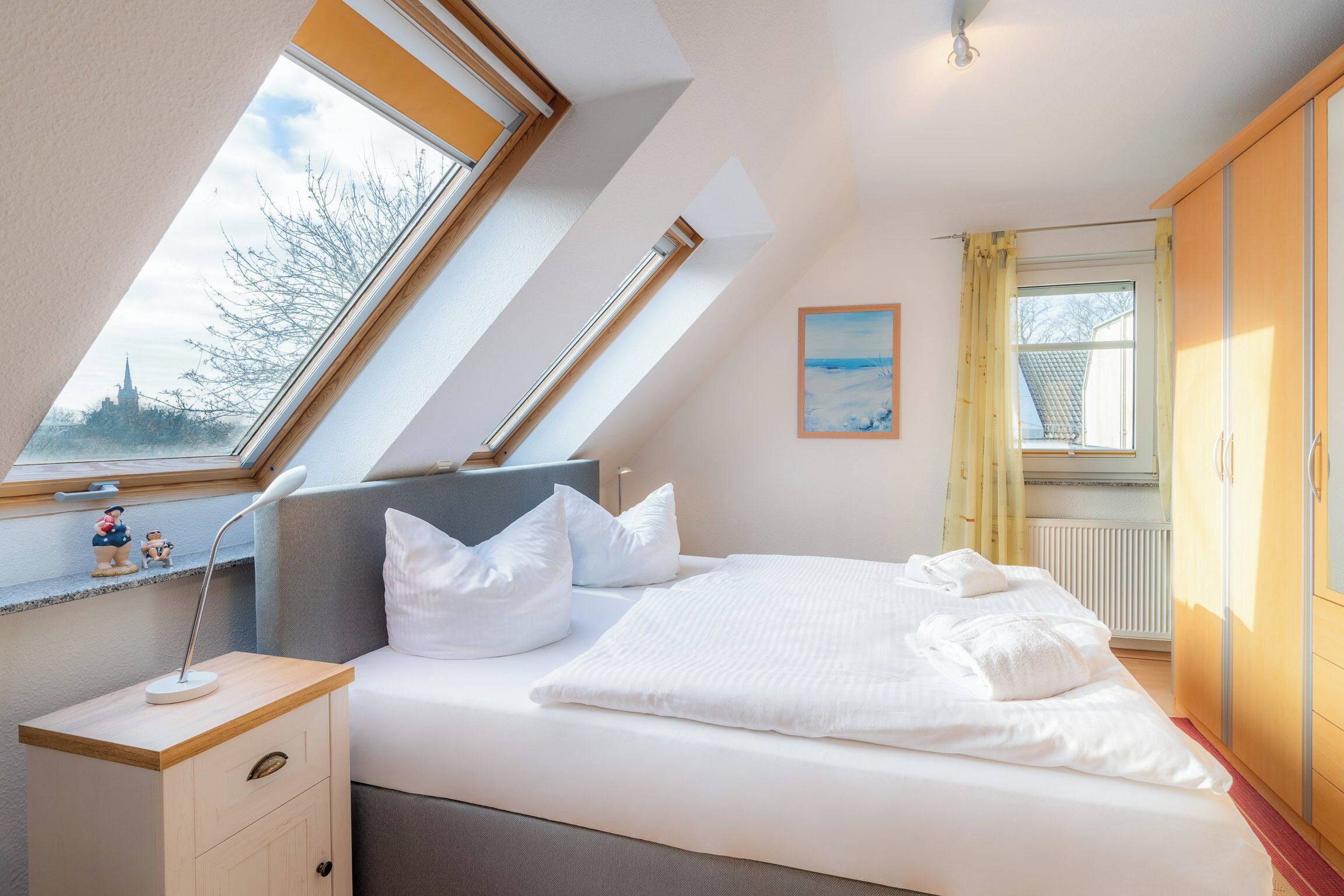 A bedroom with a skylight, a bed with white bedding, a bedside table with a lamp, and a window with curtains.