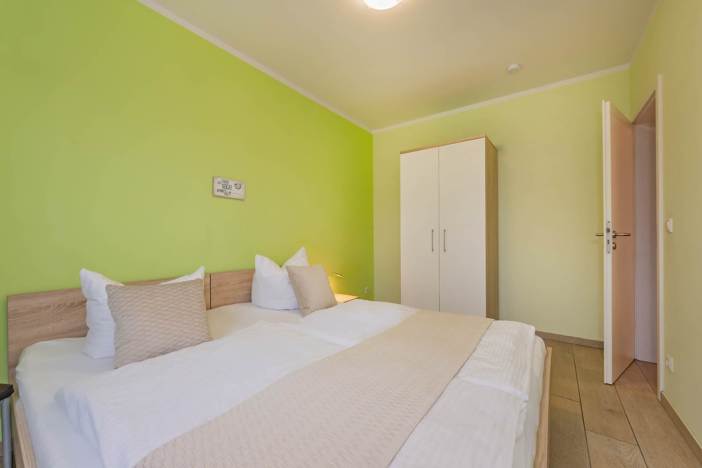 A neatly made bed with white linens and beige pillows, a white wardrobe, and a green wall.
