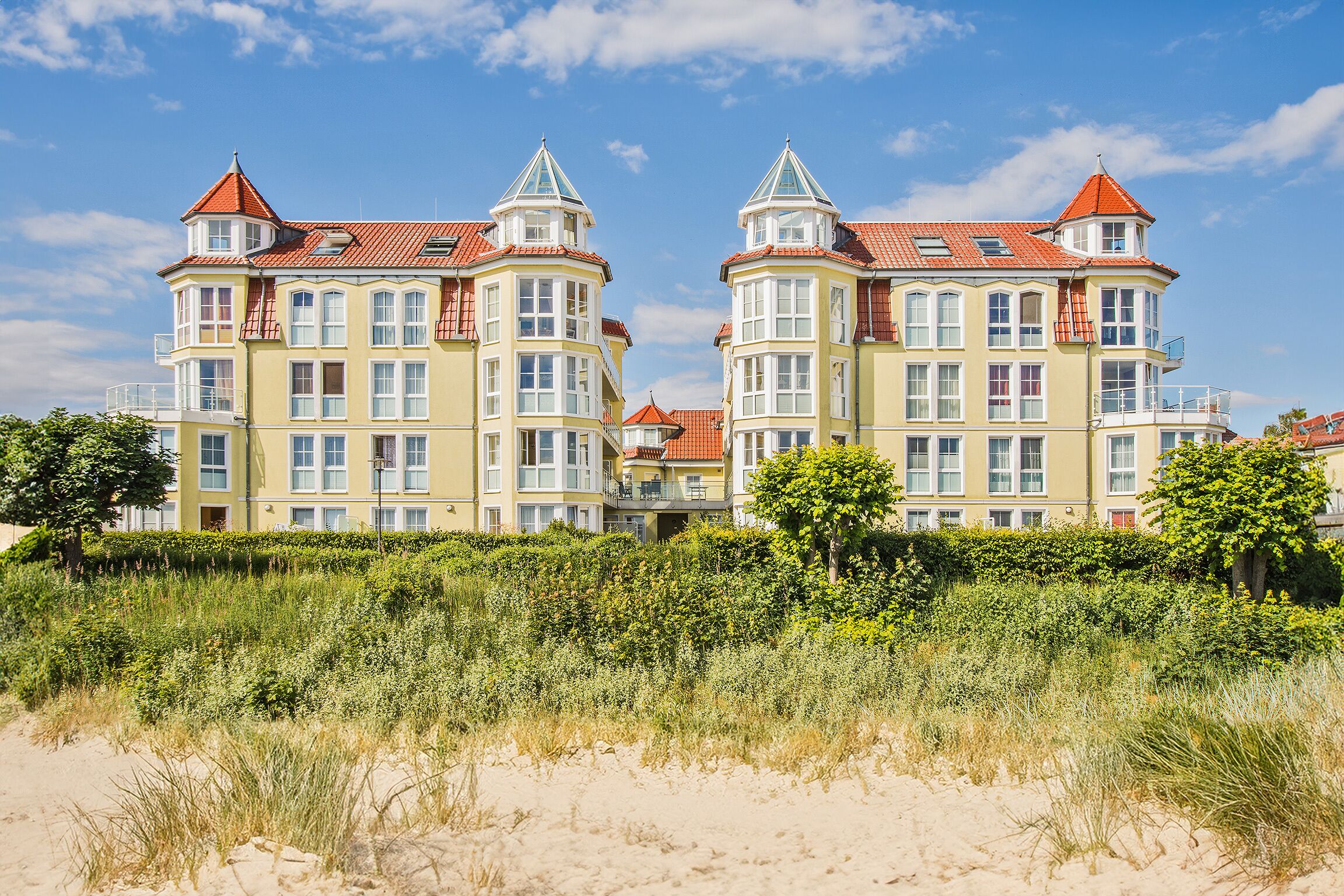 A large, yellow building with red roofs and multiple balconies, situated near a sandy beach with dense greenery in the foreground.