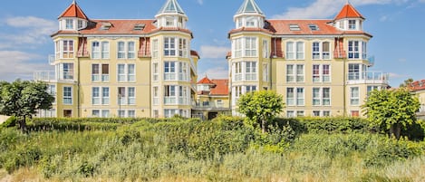 A large, yellow building with red roofs and multiple balconies, situated near a sandy beach with dense greenery in the foreground.