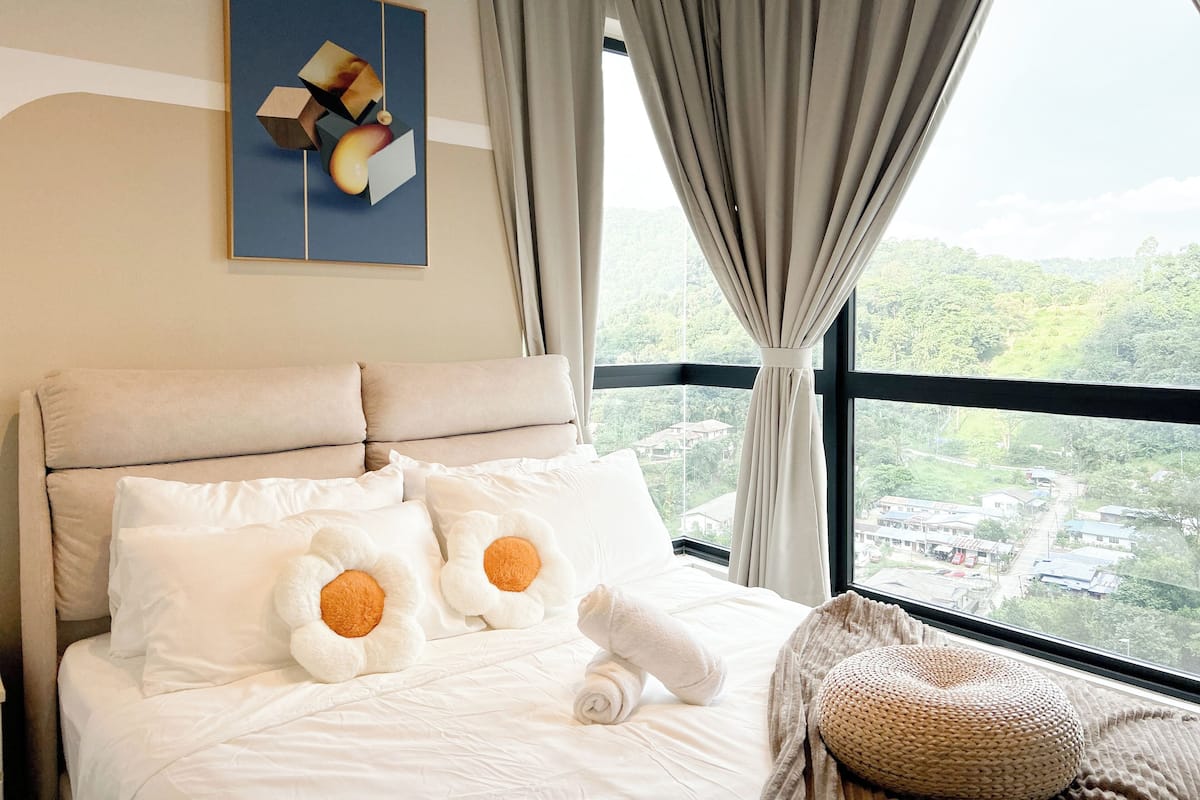 A hotel room with a bed, pillows, a window with curtains, and a painting on the wall.