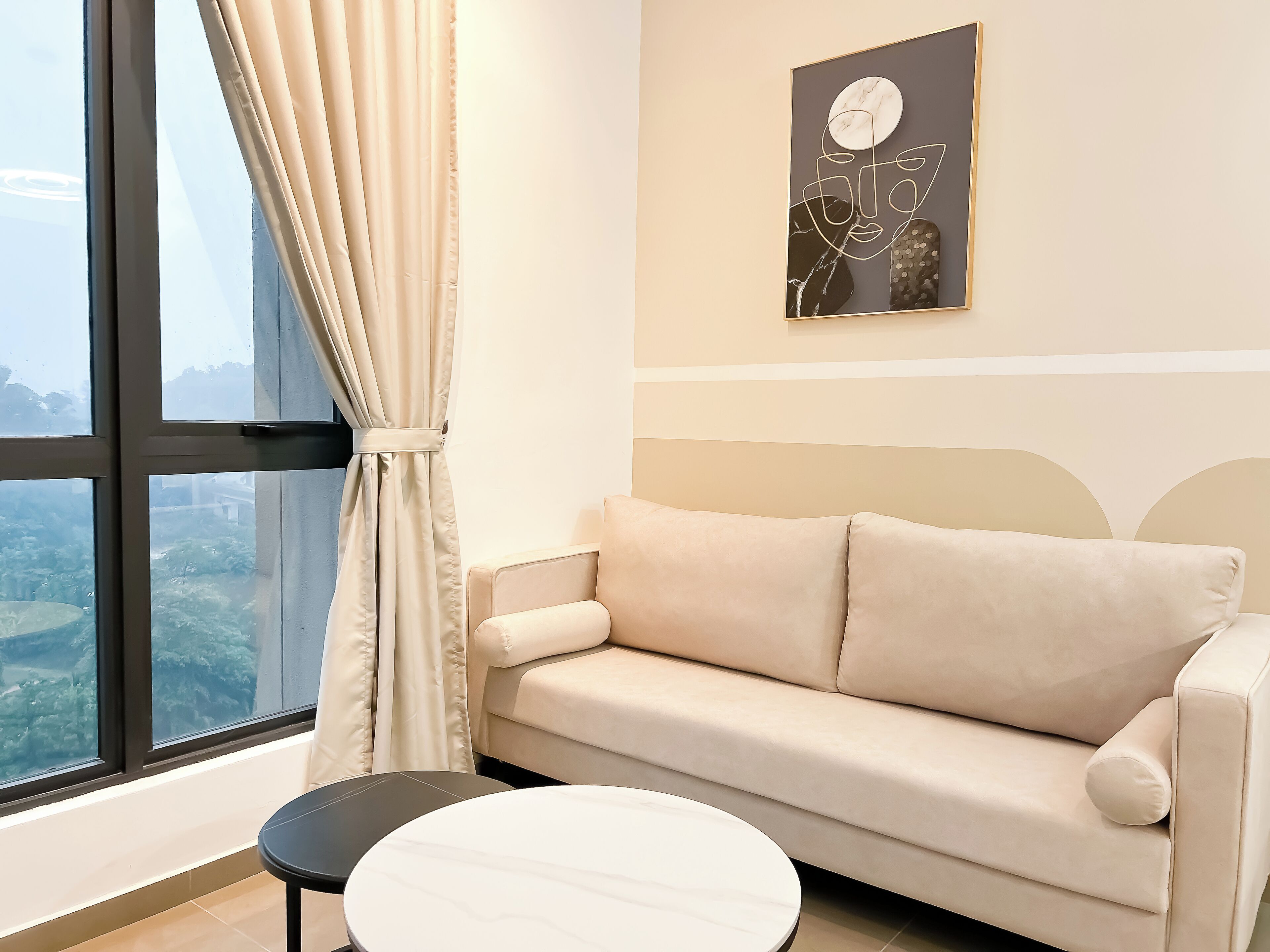 A modern living room with a beige sofa, a round white table, and a large window with curtains.