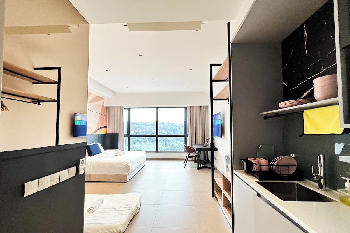 A modern hotel room with a bed, a desk, a chair, a TV, and a kitchen area.
