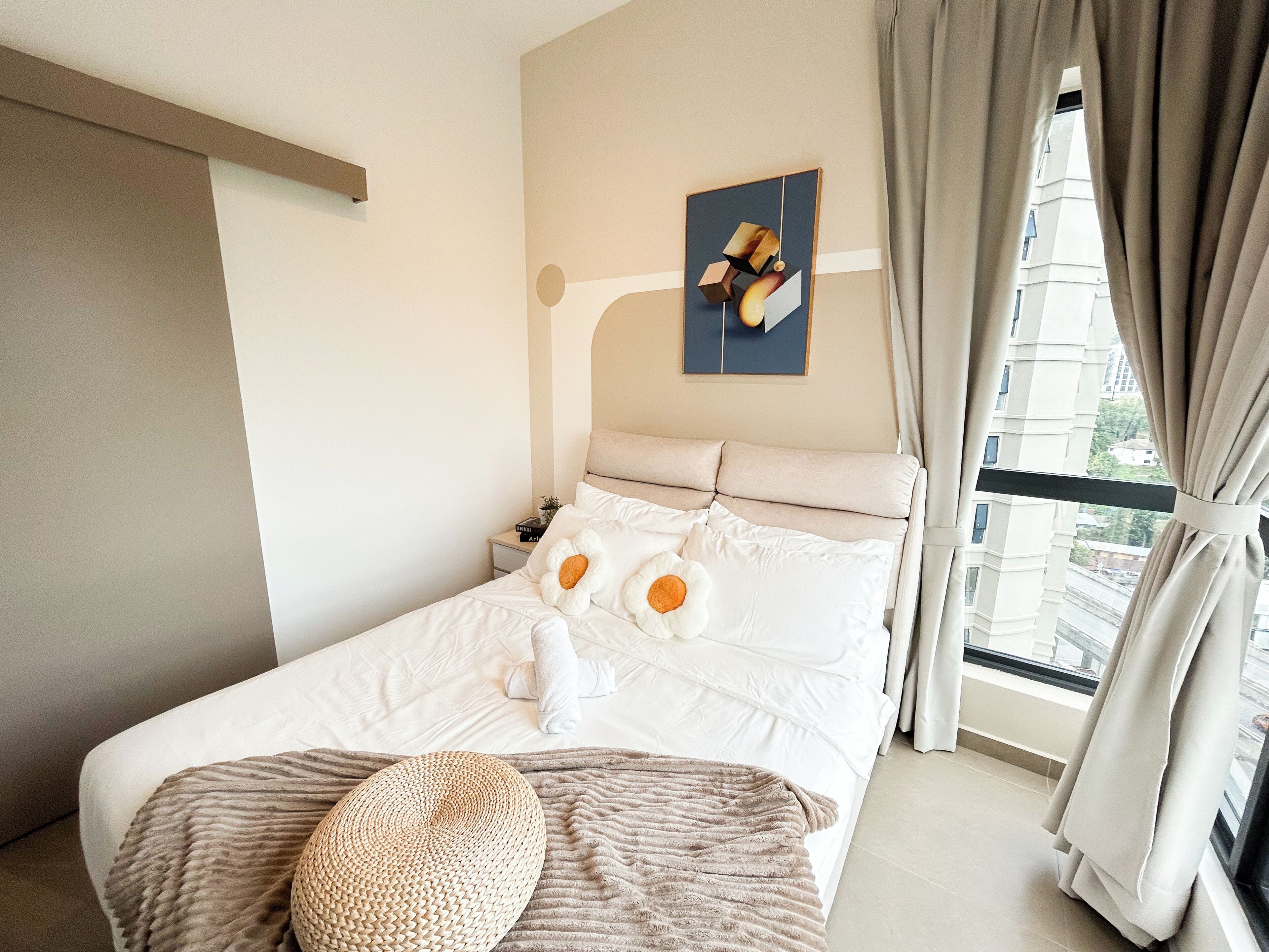 Una cama bien hecha con un cojín con forma de flor, un otoman tejido y un cuadro en la pared.