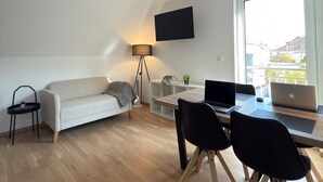 A modern living room with a sofa, a dining table with chairs, a TV mounted on the wall, and a large window providing a view of the outside. - New! Exclusive city apartment - washing machine (Karlsruhe)