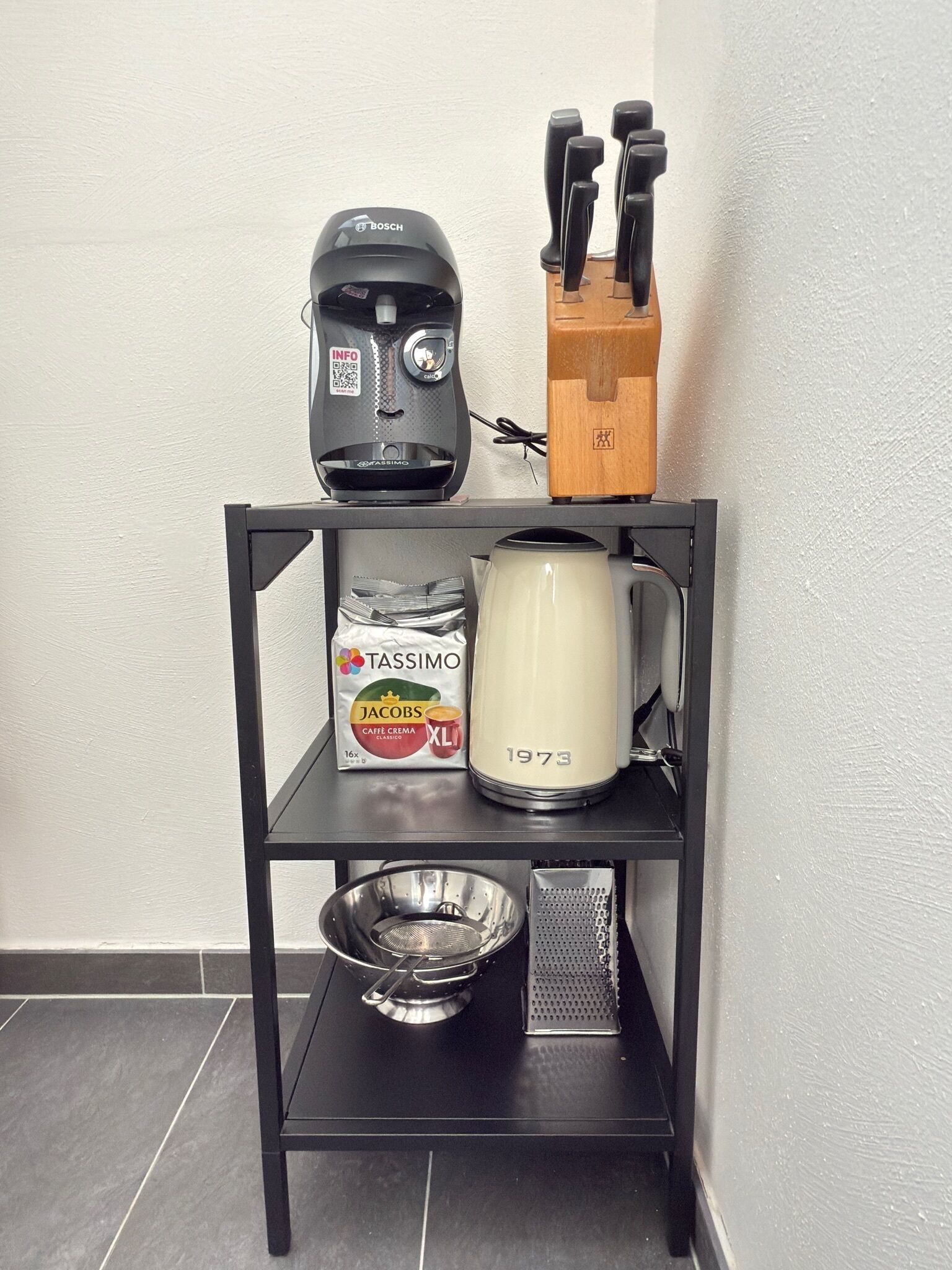 Une étagère de cuisine avec une machine à café, une bouilloire, une boîte de capsules de café Tassimo, une râpe et un saladier en métal.