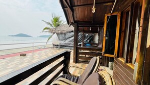 A wooden deck with a thatched roof, a wicker chair, and a view of the sea. - Palolem Luxurious Cottages (Canacona)