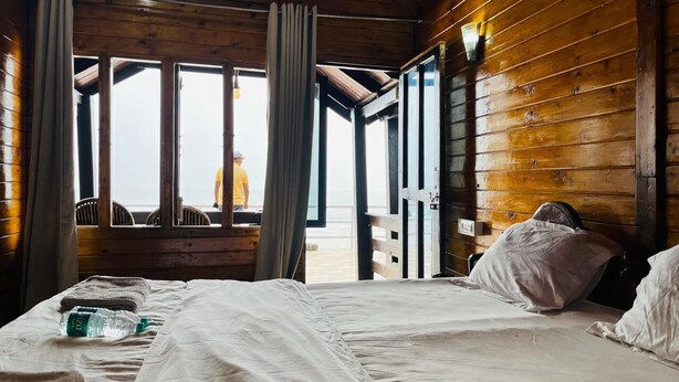A wooden cabin room with a bed, a bottle of water, and a view of the beach through large windows. - Palolem Luxurious Cottages (Canacona)