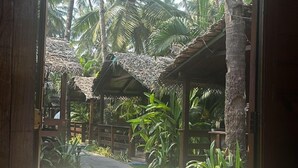 A wooden structure with a thatched roof, leading to a paved pathway surrounded by lush greenery and palm trees. - Palolem Luxurious Cottages (Canacona)