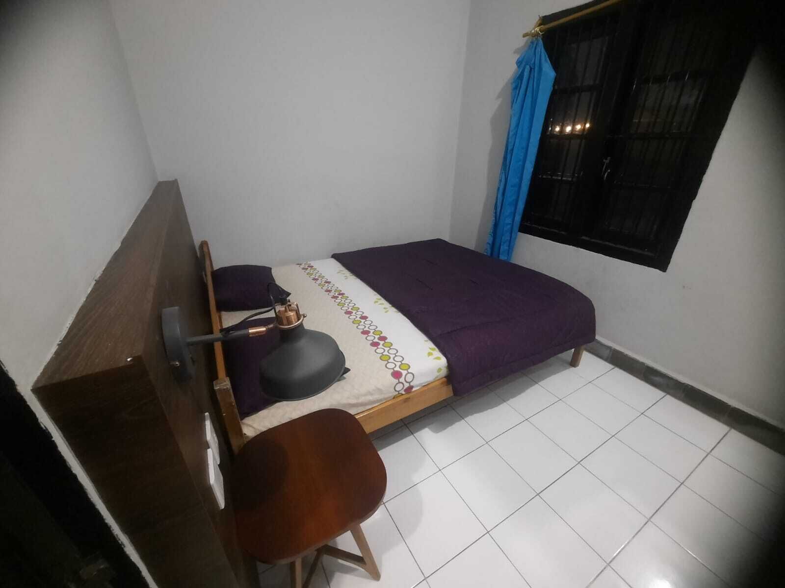 Una habitación pequeña con una cama individual, cabecera de madera, una mesita y una silla. Se aprecia una ventana con cortinas azules y una reja de metal negro.