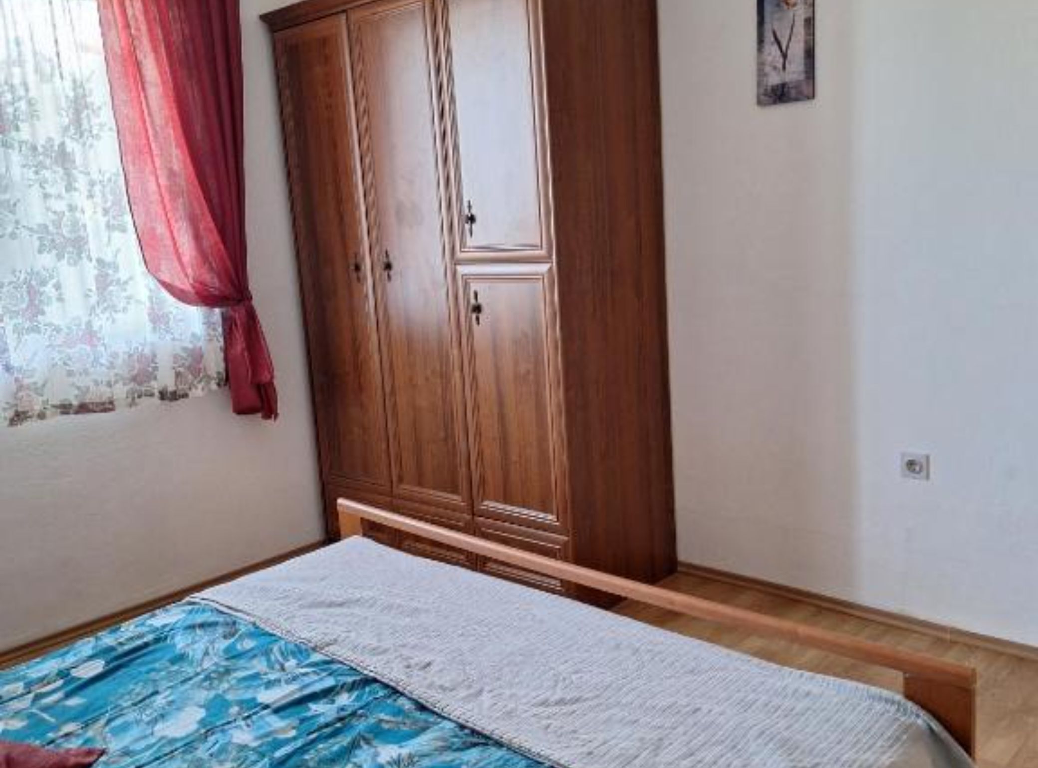 A bedroom with a wooden wardrobe, a bed with a blue and white patterned bedspread, and a window with a red curtain.