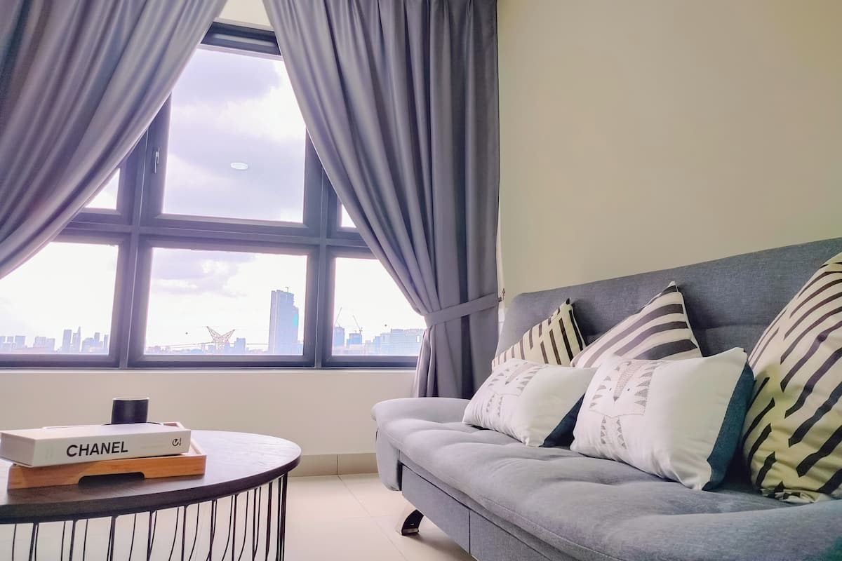 Un salon moderne avec un canapé gris, des coussins à rayures et une table basse ronde. Une boîte Chanel est posée sur la table.