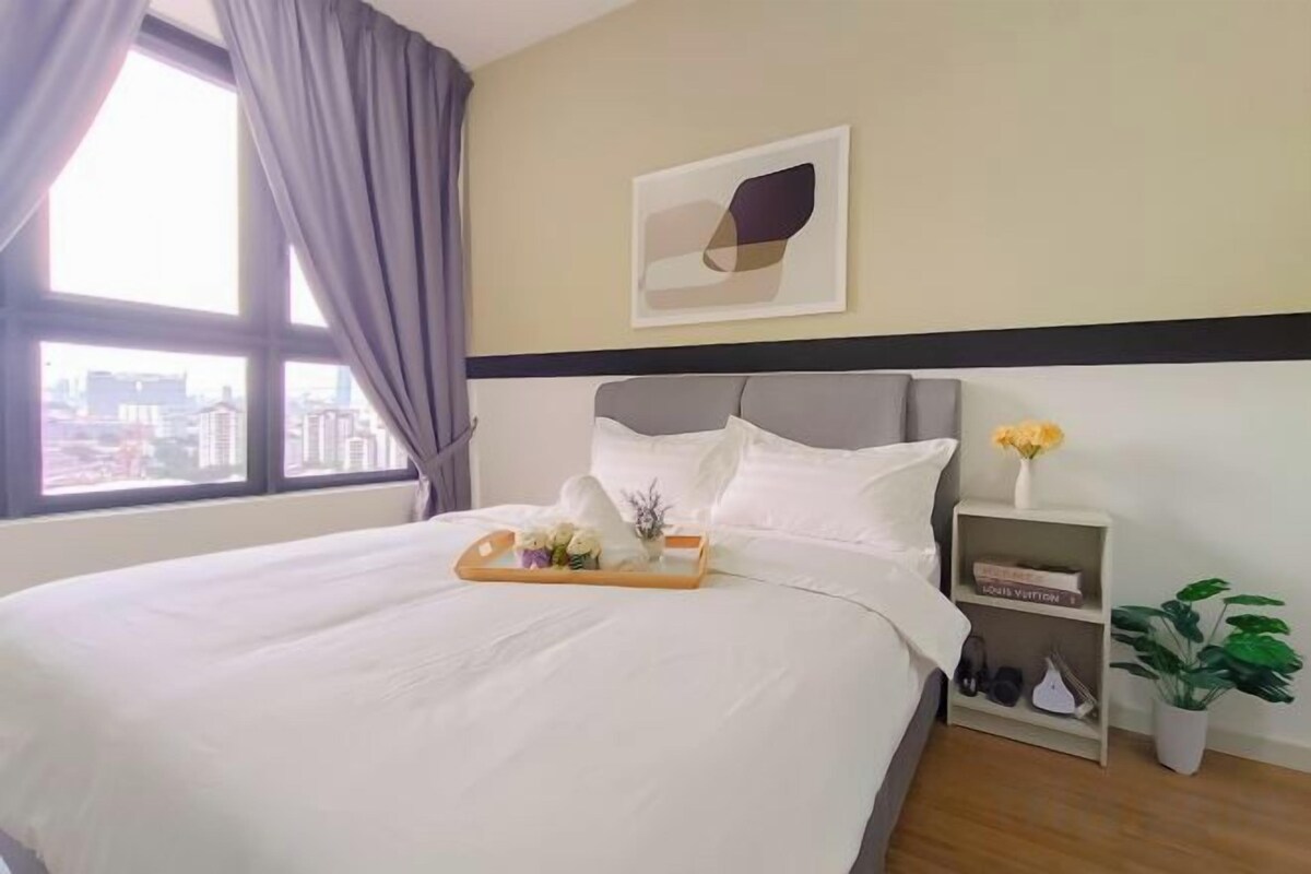 Una habitación de hotel con una cama grande, una mesita de noche con un jarrón de flores y vistas a la ciudad a través de la ventana.
