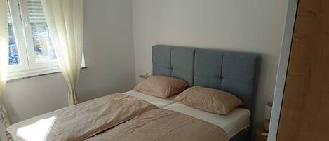 Un lit bien fait, avec une tête de lit grise, une literie beige et une fenêtre agrémentée de rideaux.