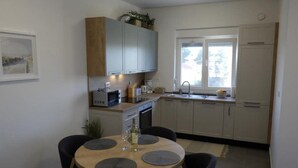 A compact kitchen with white cabinets, a sink, and a window. There is a dining table with four chairs and a bottle of wine. - Cozy Apartment Bili (Privlaka)