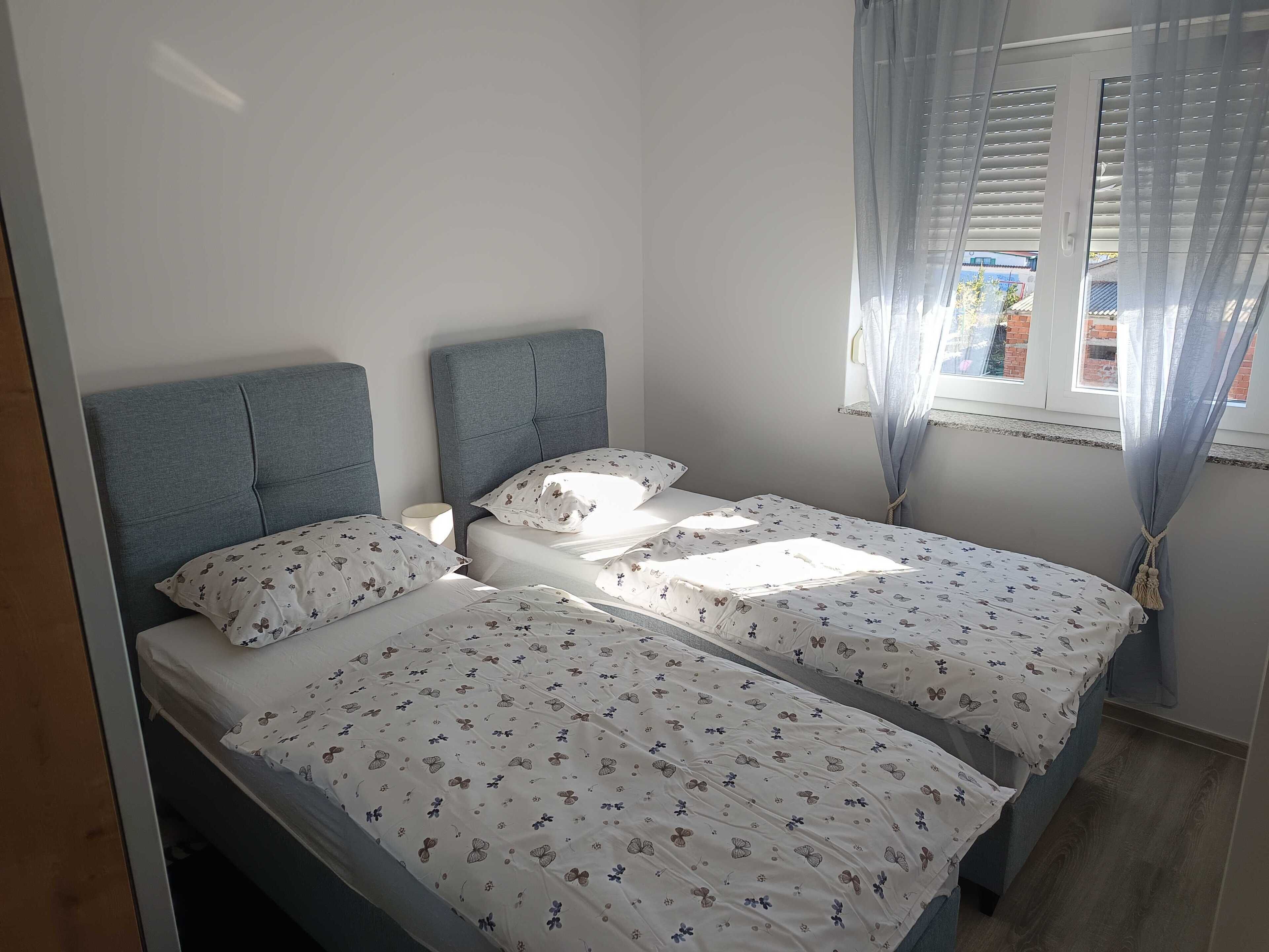 Un dormitorio con dos camas individuales, cabeceros grises, ropa de cama floral, una mesita de noche y una ventana con cortinas.