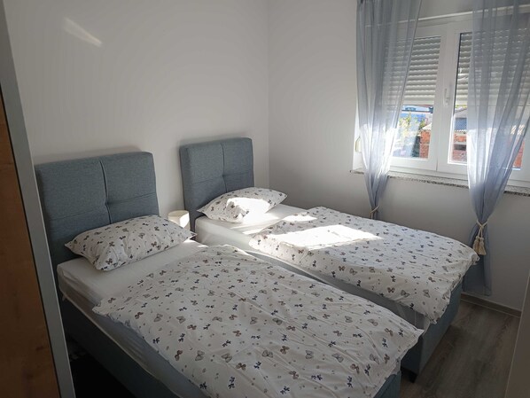 A bedroom with two single beds, a grey headboard, floral bedding, a bedside table, and a window with curtains.