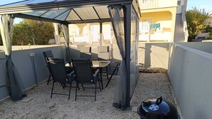 A covered patio area with a table and chairs, a grill, and a view of a building in the background. - Cozy Apartment Bili (Privlaka)