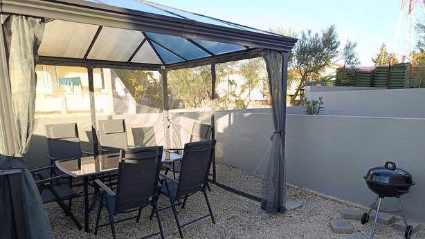An outdoor patio with a glass enclosure, a table with chairs, and a barbecue grill. - Cozy Apartment Bili (Privlaka)