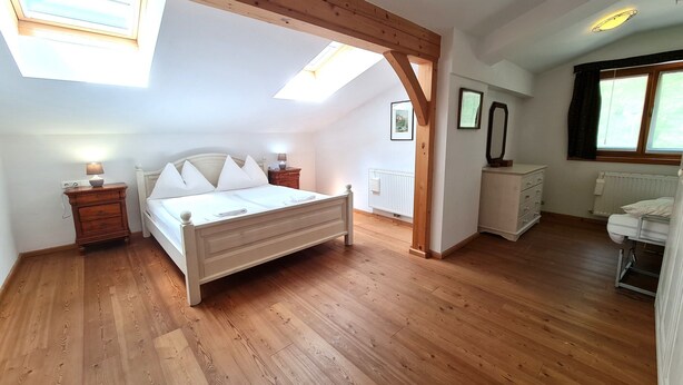 Een slaapkamer met een houten vloer, een bed met wit beddengoed, een nachtkastje met een lamp, een radiator en een raam met gordijnen.